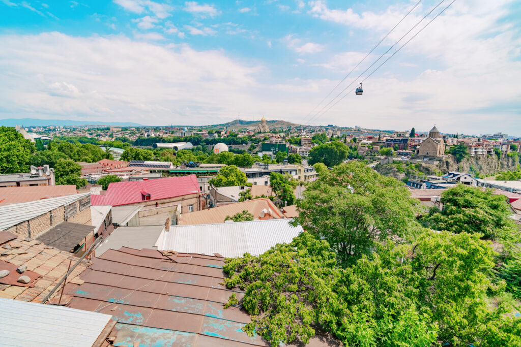 Panorama über Tiflis mit Altstadtdächern, Seilbahnkabine, Metekhi-Kirche auf Felsen und Kura-Fluss, heller Sommertag.