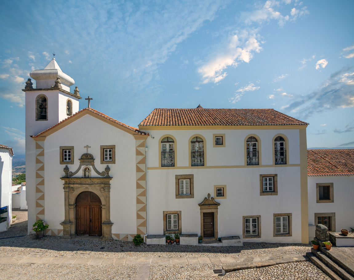 Weiße Kirche mit Glockenturm und angrenzendem Gebäude in Marvão, Kopfsteinpflasterplatz, blauer Himmel.