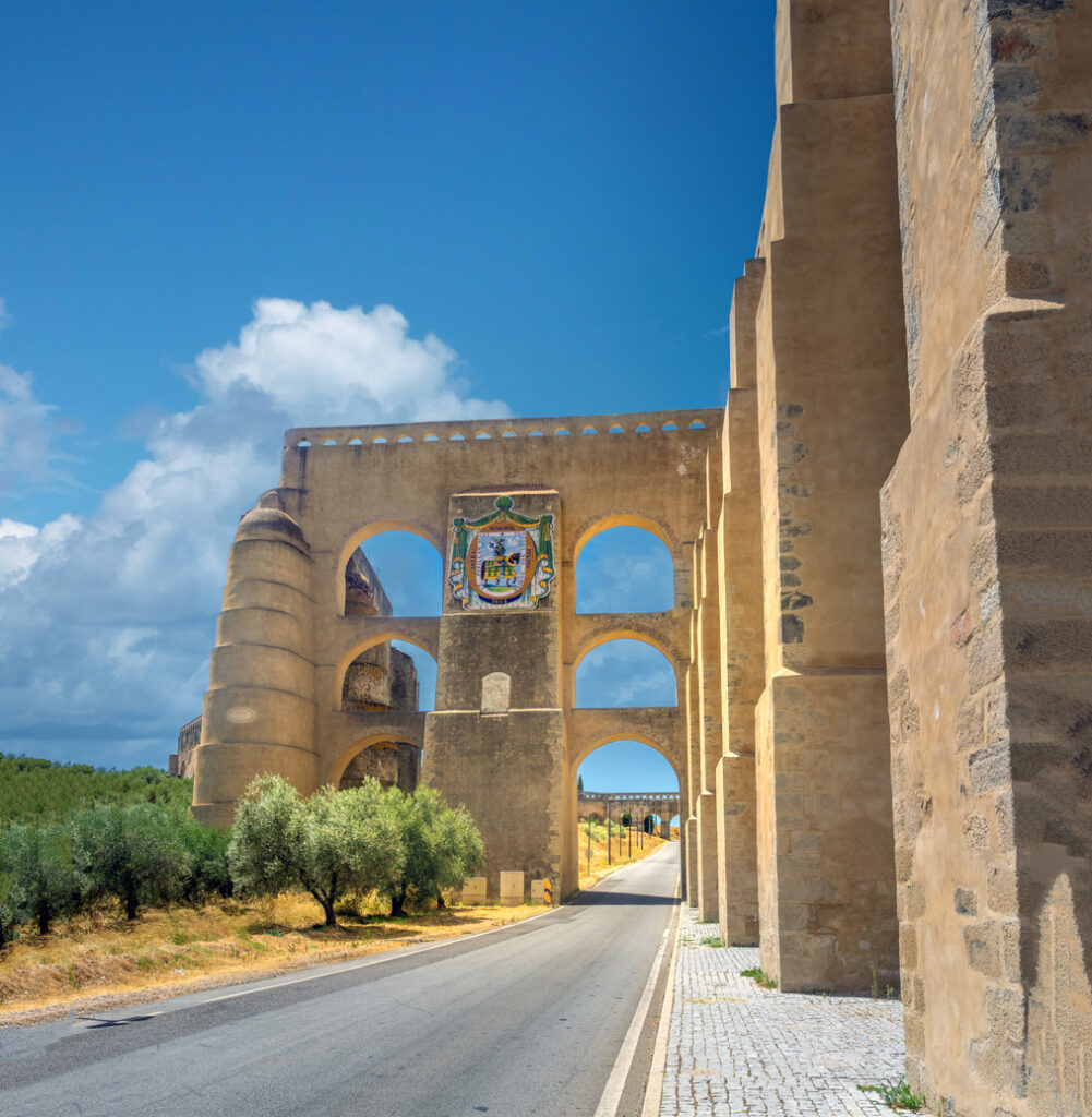 Mehrstöckiges historisches Aquädukt in Elvas, Alentejo, mit Wappen über einer Straßen­durchfahrt und Olivenbäumen am Rand