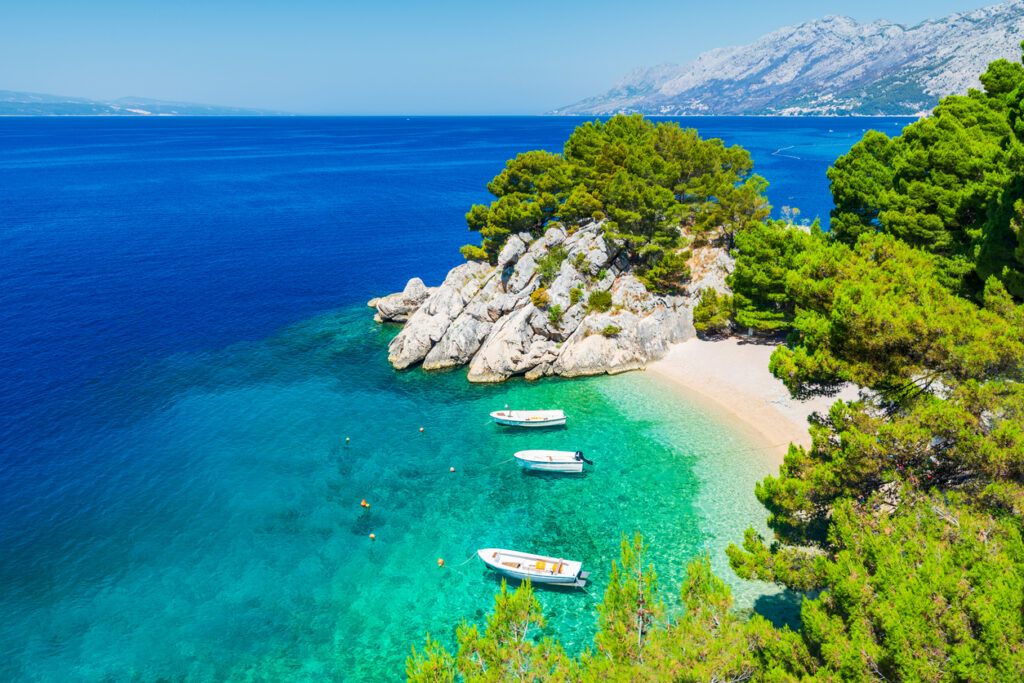 Kiesstrand Punta Rata in Brela, Kroatien – türkisblaues Wasser, Pinien und Felsvorsprung mit kleinen Booten vor dem Biokovo-Massiv