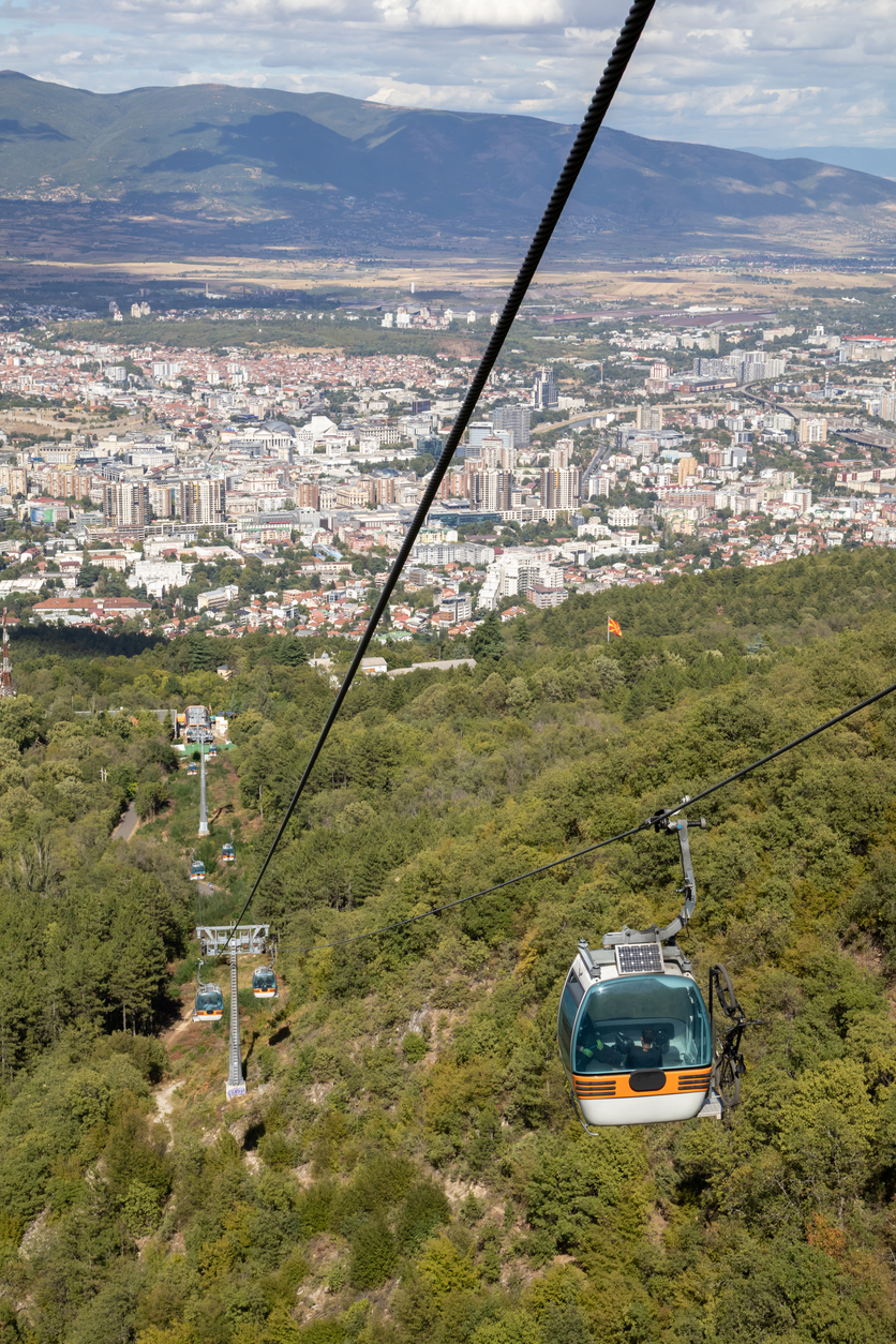 Gondel der Vodno-Seilbahn schwebt über bewaldeten Hängen; unten Skopje mit dichtem Stadtbild, dahinter Bergketten unter locker bewölktem Himmel.