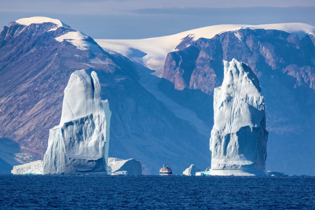 Zwei große Eisberge vor schneebedeckten Bergen; kleines Schiff fährt dazwischen.