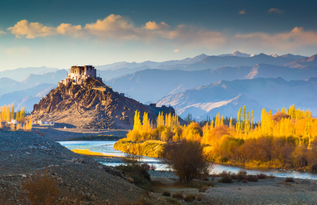 Buddhistisches Kloster auf einem Hügel in Ladakh, Indien, umgeben von bunten Bäumen und den Bergen des Himalaya.