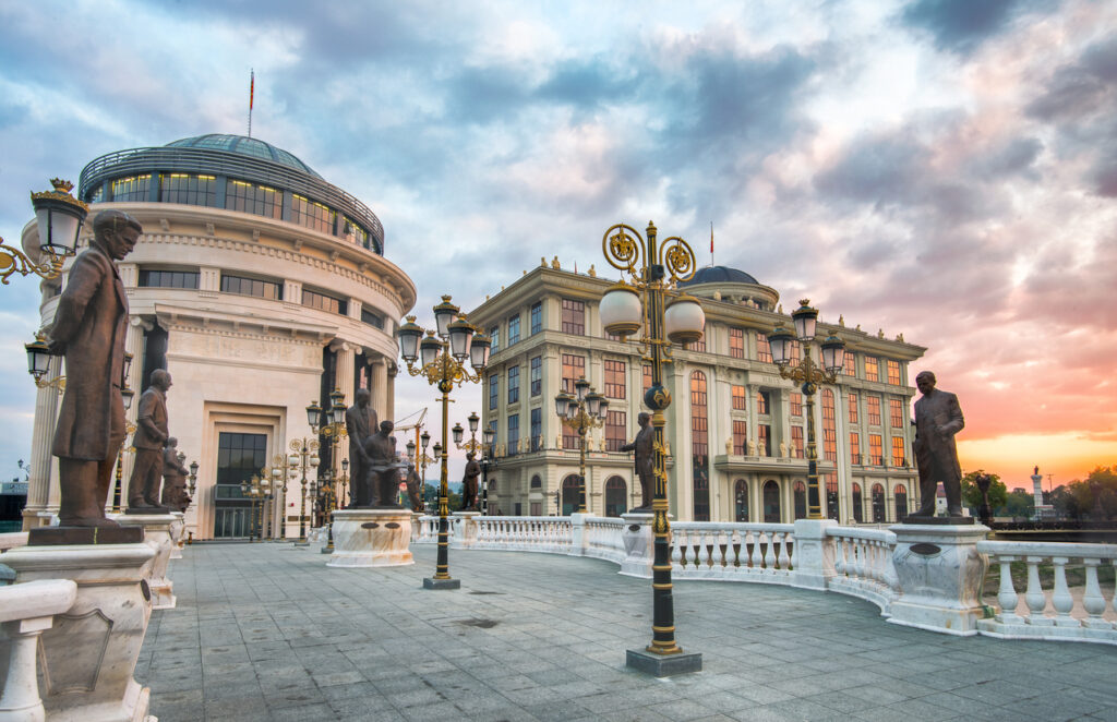 Fußgängerbrücke in Skopje mit zahlreichen Bronzestatuen und verzierten Laternen, dahinter kuppelgekrönte Museums- und Regierungsgebäude am Fluss Vardar im warmen Abendlicht.