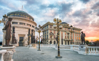 Fußgängerbrücke in Skopje mit zahlreichen Bronzestatuen und verzierten Laternen, dahinter kuppelgekrönte Museums- und Regierungsgebäude am Fluss Vardar im warmen Abendlicht.
