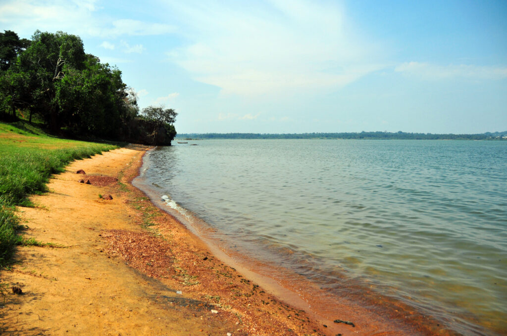 Rötlicher Sandstrand am Victoriasee, flaches Wasser, Bäume und blauer Himmel.