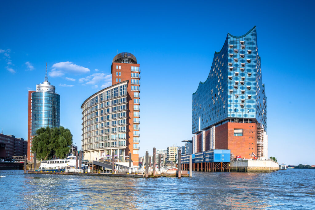 Hamburg, Elbphilharmonie an der Elbe mit gläsernem Wellendach auf rotem Backsteinsockel, links moderne HafenCity-Bauten, Wasser im Vordergrund, klarer Himmel