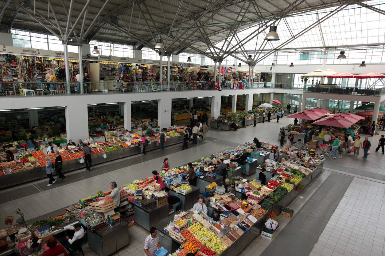 Innenansicht der modernen, lichtdurchfluteten Markthalle in Podgorica mit zahlreichen Verkaufsständen für frisches Obst, Gemüse, Käse und Blumen sowie vielen Besuchern.