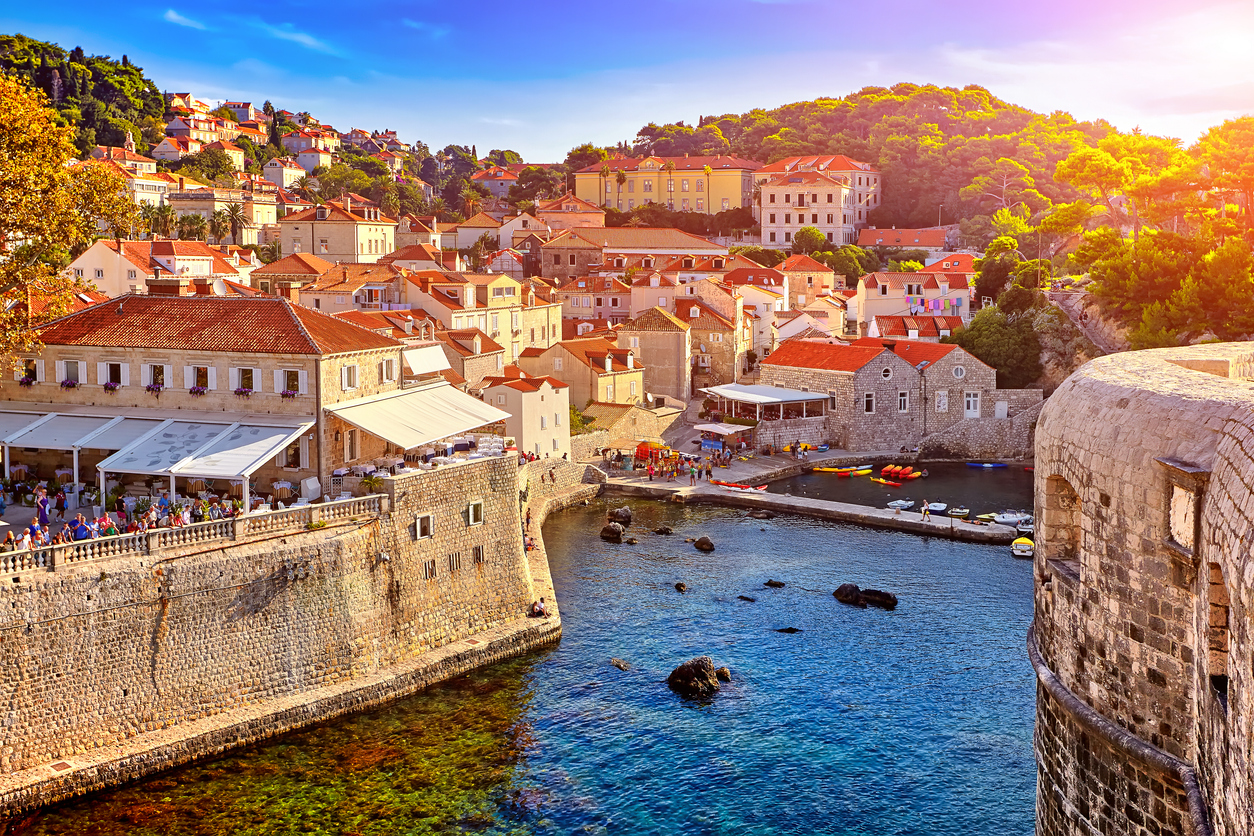 Blick über Dubrovniks Stadtmauern auf Altstadthäuser mit roten Dächern und kleinem Hafen am türkisblauen Meer.