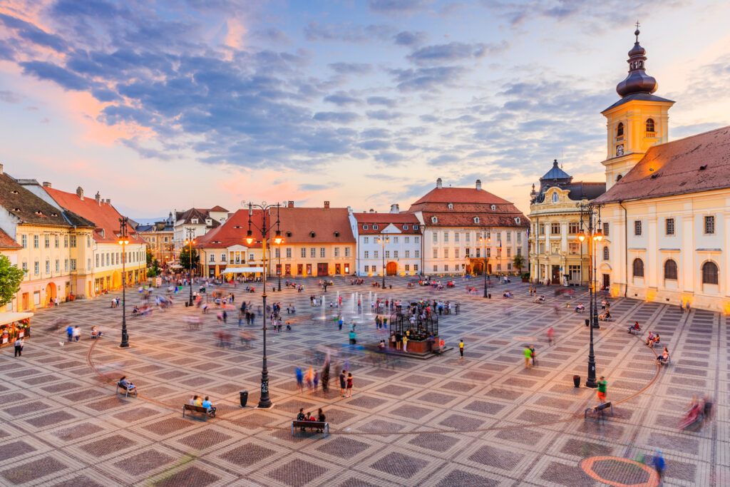Der zentrale Marktplatz von Sibiu in Rumänien bei Sonnenuntergang, gesäumt von historischen Gebäuden und Menschen, die flanieren und verweilen.