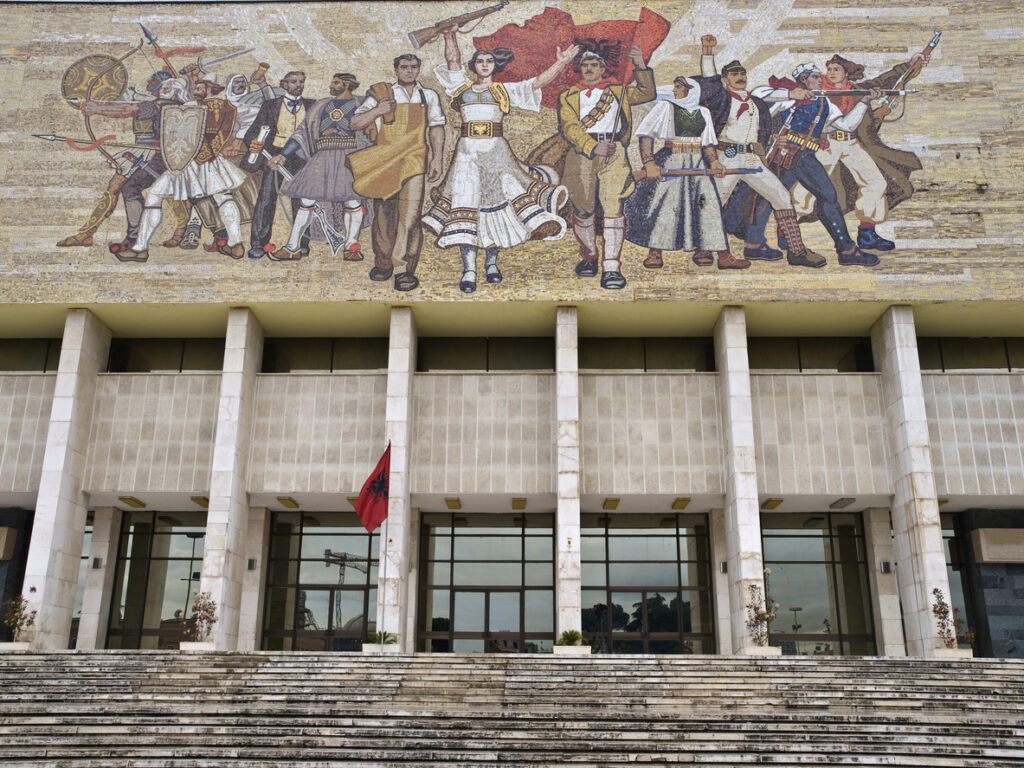 Frontalansicht des Nationalhistorischen Museums in Tirana: breite Treppe, albanische Flagge und großes Fassadenmosaik „Die Albaner“ über der Säulenfassade