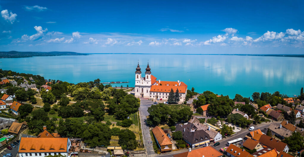 Luftaufnahme von Tihany mit der barocken Benediktinerabtei (Doppelturm), roten Ziegeldächern und dem türkisblauen Balaton im Hintergrund.