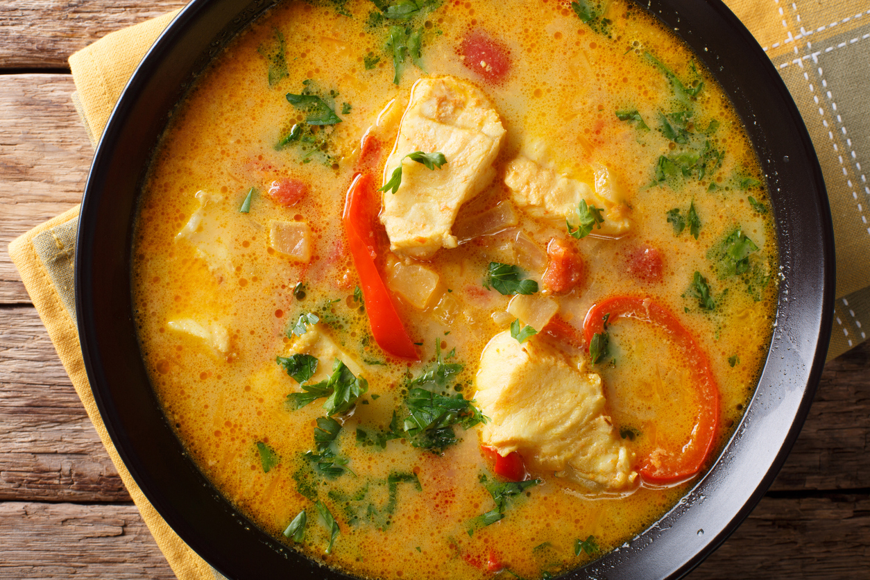 Teller mit Moqueca baiana zeigt weiße Fischstücke in gelblicher Kokossauce mit roten Paprikastreifen und frischen Kräutern.