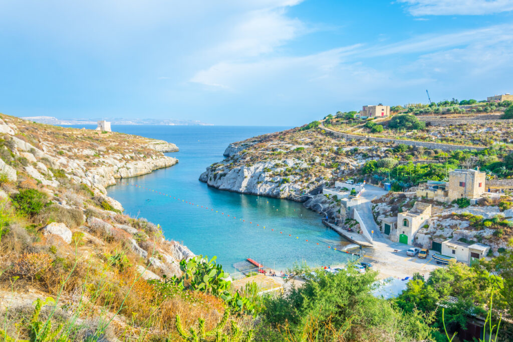 Schmale Felsbucht mit türkisblauem Wasser, wenigen Häusern und einem Küstenwachturm auf Gozo.