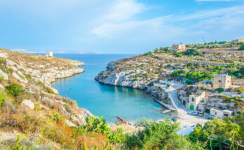 Schmale Felsbucht mit türkisblauem Wasser, wenigen Häusern und einem Küstenwachturm auf Gozo.
