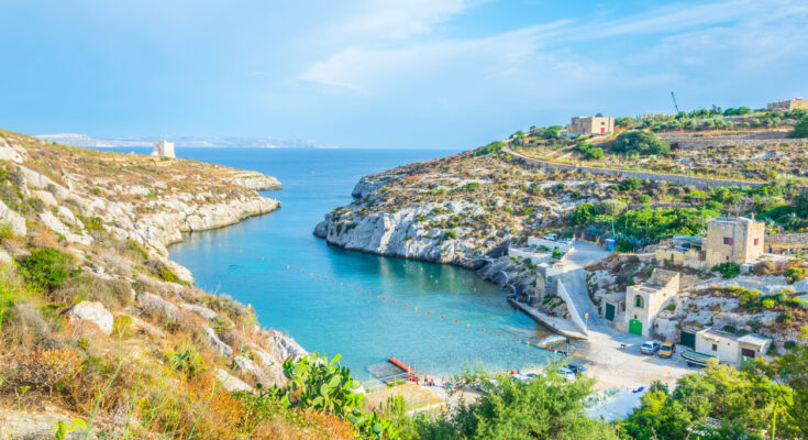 Schmale Felsbucht mit türkisblauem Wasser, wenigen Häusern und einem Küstenwachturm auf Gozo.