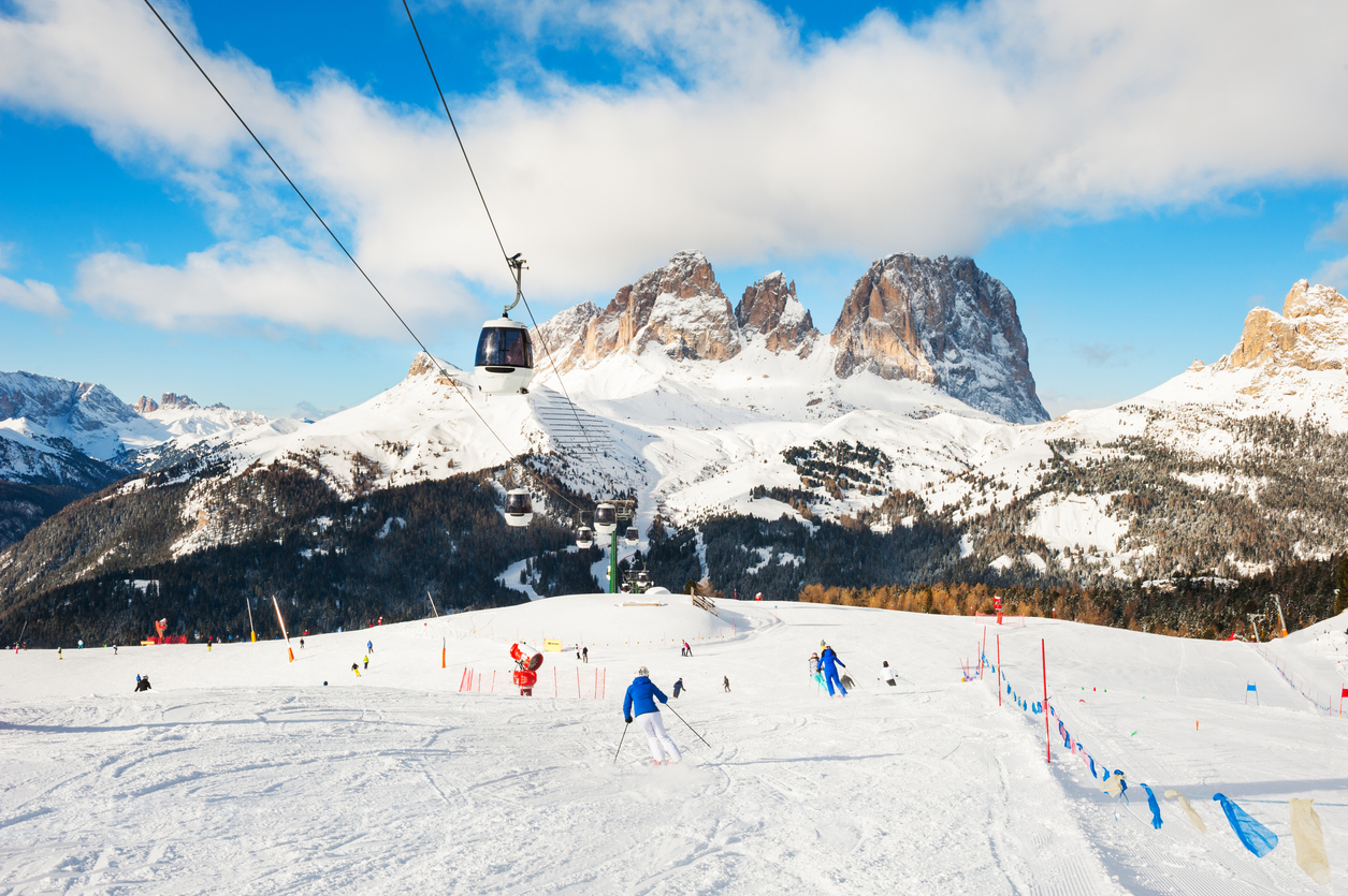 Breite Piste bei Canazei mit Gondelbahn, Skifahrern und gezackten Dolomiten.