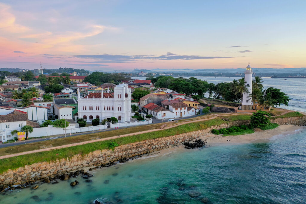 Luftaufnahme des UNESCO-Galle-Fort mit weißem Leuchtturm, Stadtmauer und Kolonialgebäuden am türkisblauen Meer bei weichem Abendlicht.