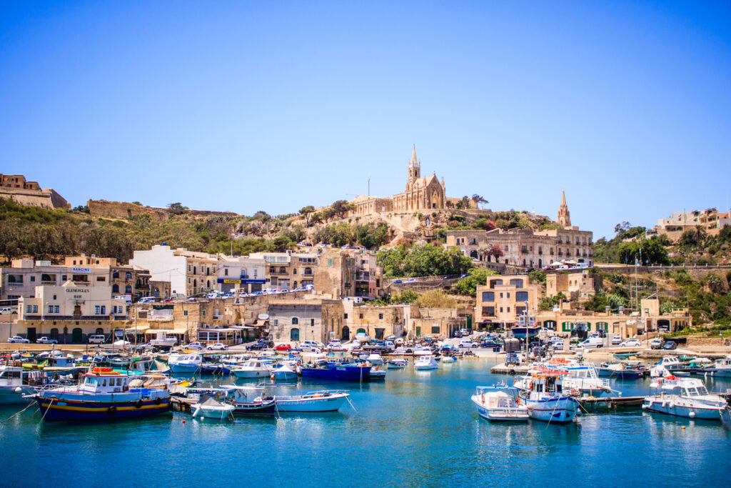Blick auf den Hafen von Mġarr auf Gozo mit Booten und der Kirche Our Lady of Lourdes auf dem Hügel.