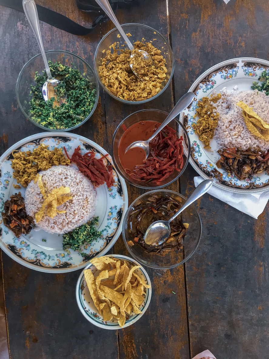 Draufsicht auf sri-lankisches Rice-and-Curry mit rotem Reis, grünen Sambols, Gemüse- und Linsencurrys sowie knusprigen Papadams in Schälchen.