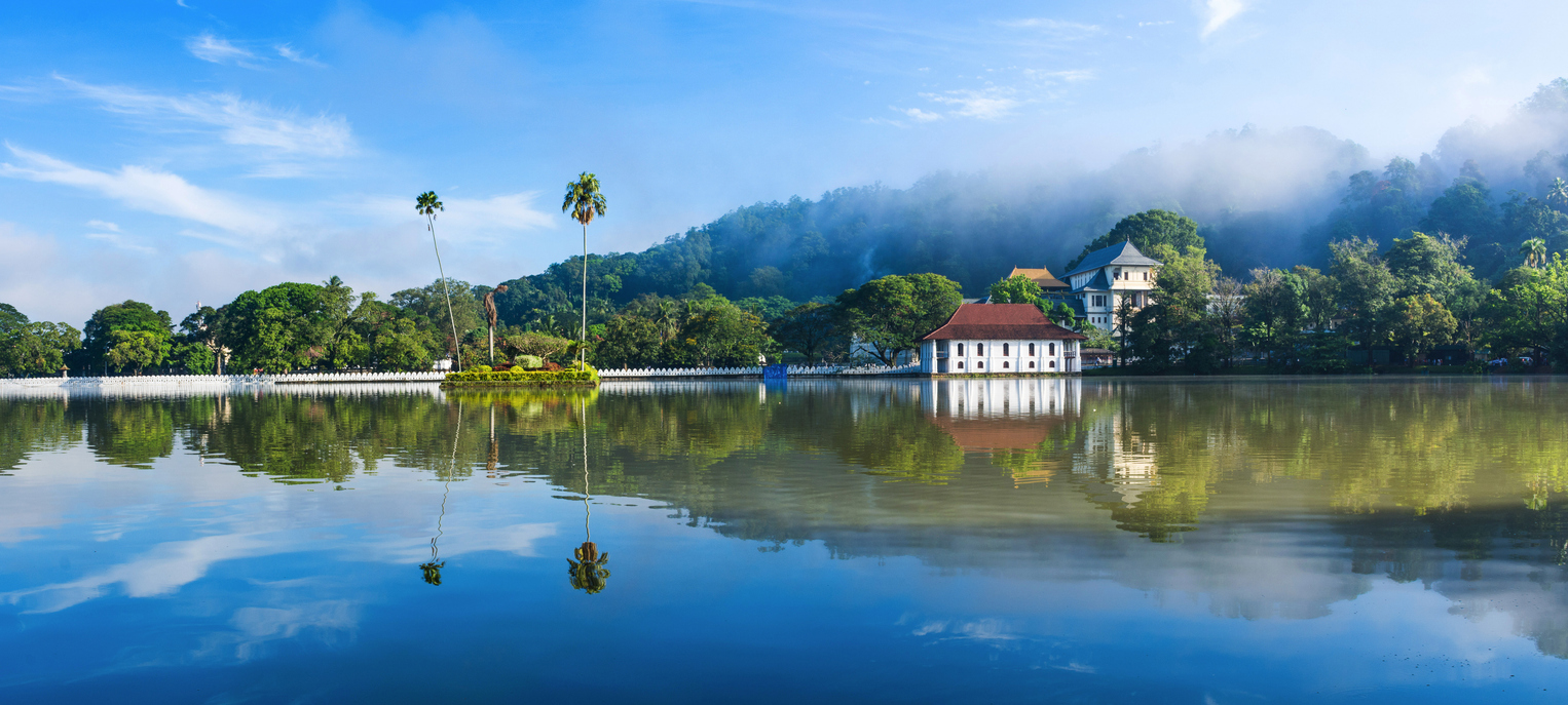 Panoramablick auf den Zahntempel Sri Dalada Maligawa am Kandy Lake mit weißen Mauern, Palmen und klarer Spiegelung im Wasser.
