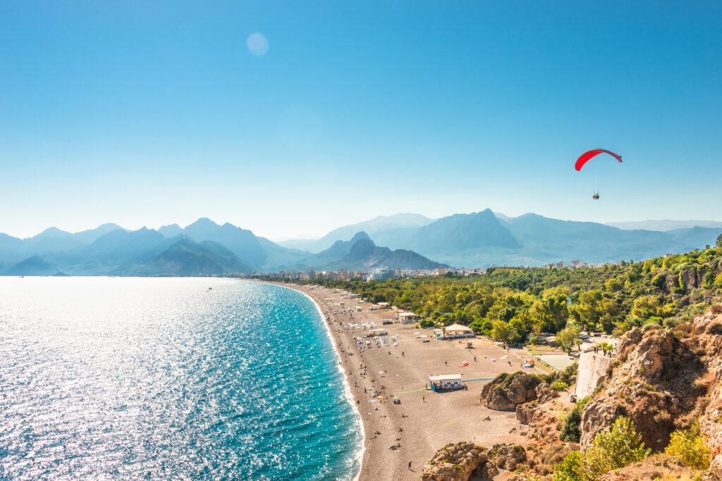 Langer Kiesstrand mit türkisfarbenem Meer und Paraglider vor den Taurusbergen.