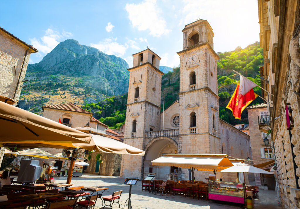 Romanische Kathedrale Sveti Tripun mit zwei Glockentürmen in der Altstadt von Kotor, Cafés und Sonnenschirmen, dahinter steile Berge.
