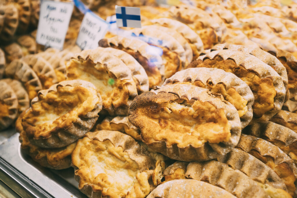 Stapel karelischer Piroggen (Karjalanpiirakka) mit gefaltetem Roggenrand und heller Reisfüllung in einer Auslage; kleines Finnland-Fähnchen.