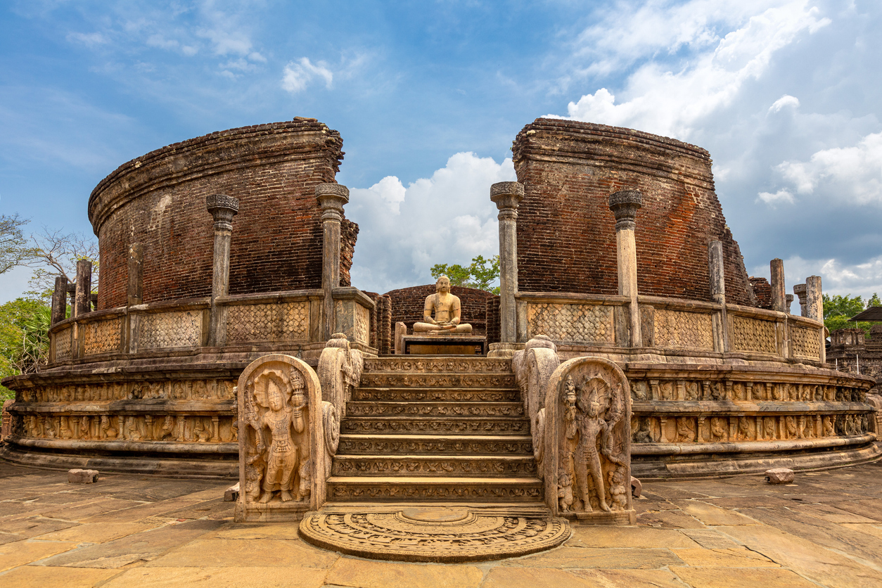 Die kreisförmige Vatadage von Polonnaruwa mit Treppe, Mondstein, Wächtersteinen und sitzendem Buddha unter blauem Himmel.
