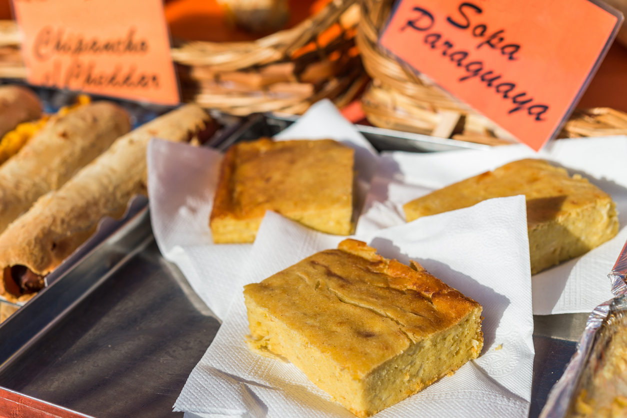 Goldbraune Stücke Sopa paraguaya liegen auf Servietten auf einem Marktstand mit Schild.