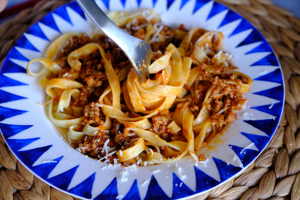 Bandnudeln mit tomatigem Hackfleischragù, Parmesanraspeln und Gabel auf blau-weißem Teller.