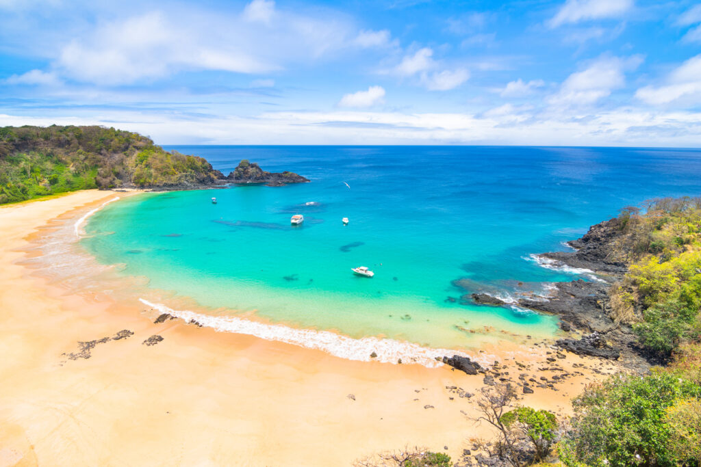 Türkisfarbene Bucht mit goldgelbem Sandstrand, kleinen Booten und Felsen auf Fernando de Noronha, Brasilien.