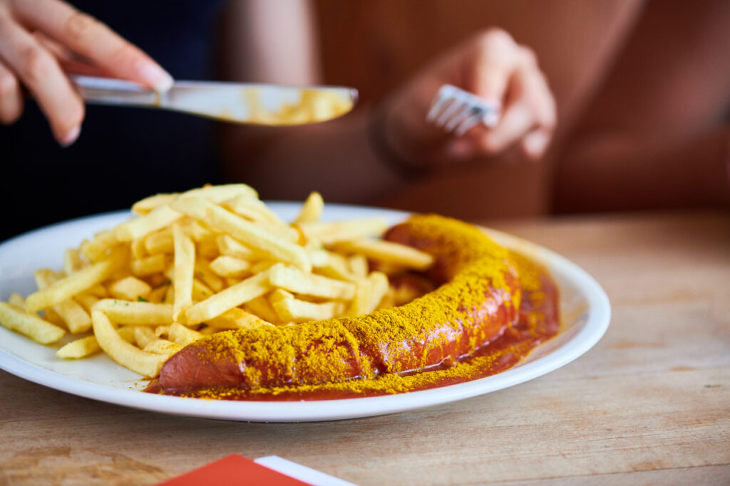 Aufgeschnittene Currywurst in Tomaten-Curry-Sauce mit Pommes auf weißem Teller.
