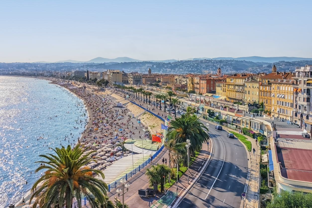 Nizza: Blick über die Promenade des Anglais mit palmengesäumter Küstenstraße, belebtem Kiesstrand und azurblauem Mittelmeer.