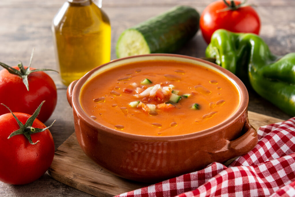 Braune Keramikschale mit orange-rotem Gazpacho, oben gewürfelte Gurke und Tomate, daneben Tomaten, Gurke, Paprika und Olivenöl.