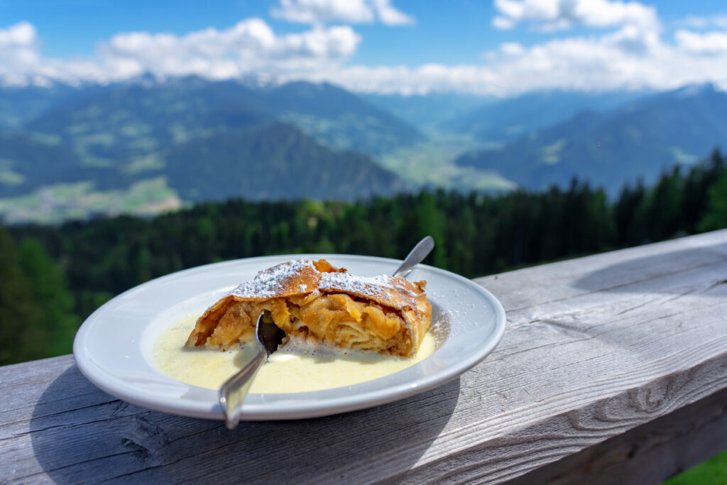Ein Stück Apfelstrudel mit Puderzucker und Vanillesauce auf einem Teller vor Bergpanorama.