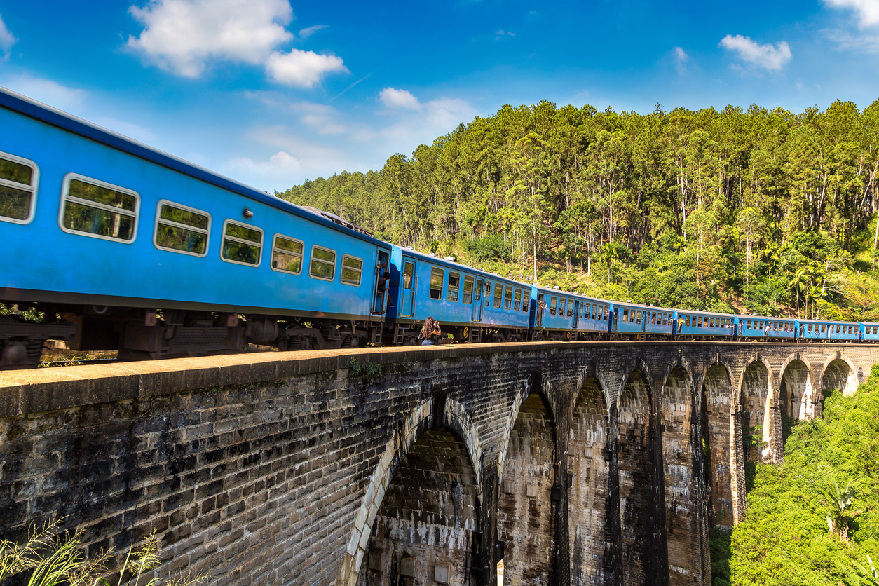 Ein blauer Zug überquert die steinerne Neun-Bogen-Brücke in Ella, Sri Lanka, umgeben von dichtem Grün.
