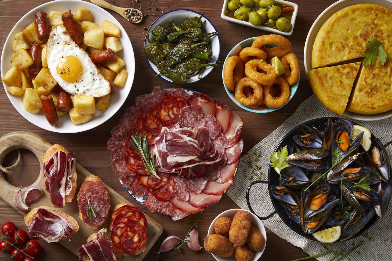 Obenansicht eines Tisches mit verschiedenen spanischen Tapas wie Tortilla, Muscheln, Calamares, Oliven, Croquetas, Wurstaufschnitt und Kartoffeln mit Ei.
