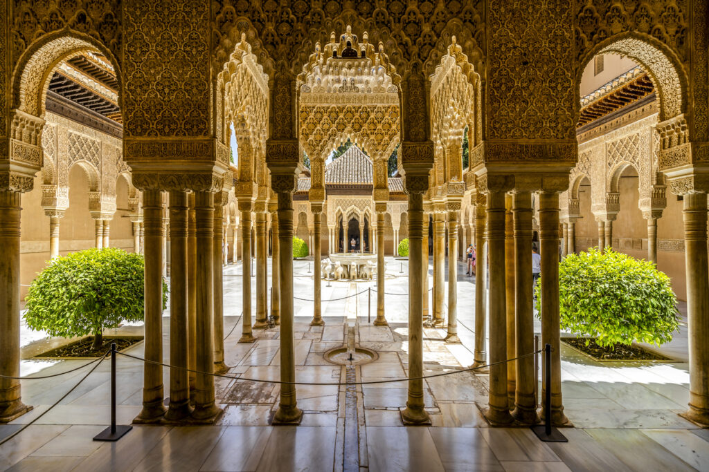 Blick durch reich verzierte Bögen und Säulen auf den Löwenhof der Alhambra in Granada.