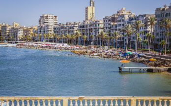 Blick auf den Stadtstrand von Alexandria am Mittelmeer mit vielen Badenden, bunten Sonnenschirmen, Palmenallee und Wohnhäusern entlang der Corniche.