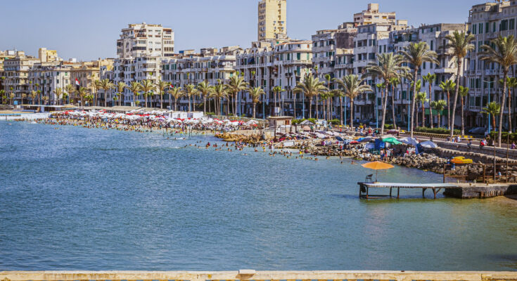 Blick auf den Stadtstrand von Alexandria am Mittelmeer mit vielen Badenden, bunten Sonnenschirmen, Palmenallee und Wohnhäusern entlang der Corniche.