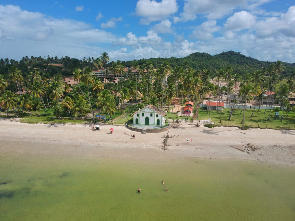 Kleine weiß-grüne Kapelle am Strand vor Palmen, ruhigem grünblauem Wasser und Badenden in Praia dos Carneiros, Pernambuco.