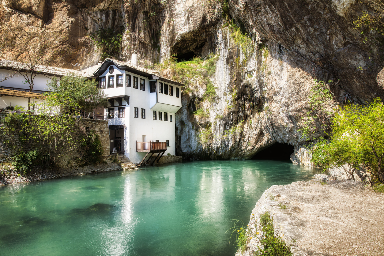 Weißes Derwischkloster am türkisfarbenen Fluss vor einer Felswand mit Höhleneingang.