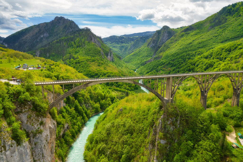 Panorama der gewaltigen Tara-Schlucht im Durmitor-Nationalpark mit der Bogenbrücke Đurđevića Tara über smaragdgrünem Fluss und bewaldeten Bergen.