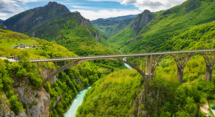Panorama der gewaltigen Tara-Schlucht im Durmitor-Nationalpark mit der Bogenbrücke Đurđevića Tara über smaragdgrünem Fluss und bewaldeten Bergen.