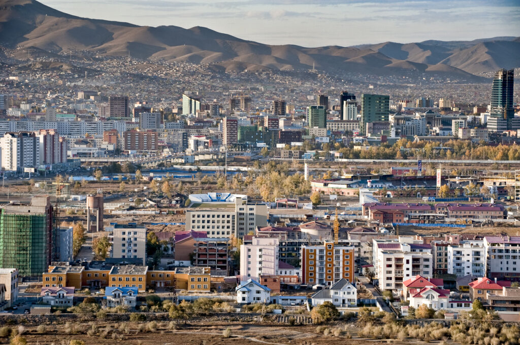 Panoramablick auf Ulaanbaatar mit Hochhäusern, Wohnvierteln und herbstlichen Bäumen vor welligen Bergketten.