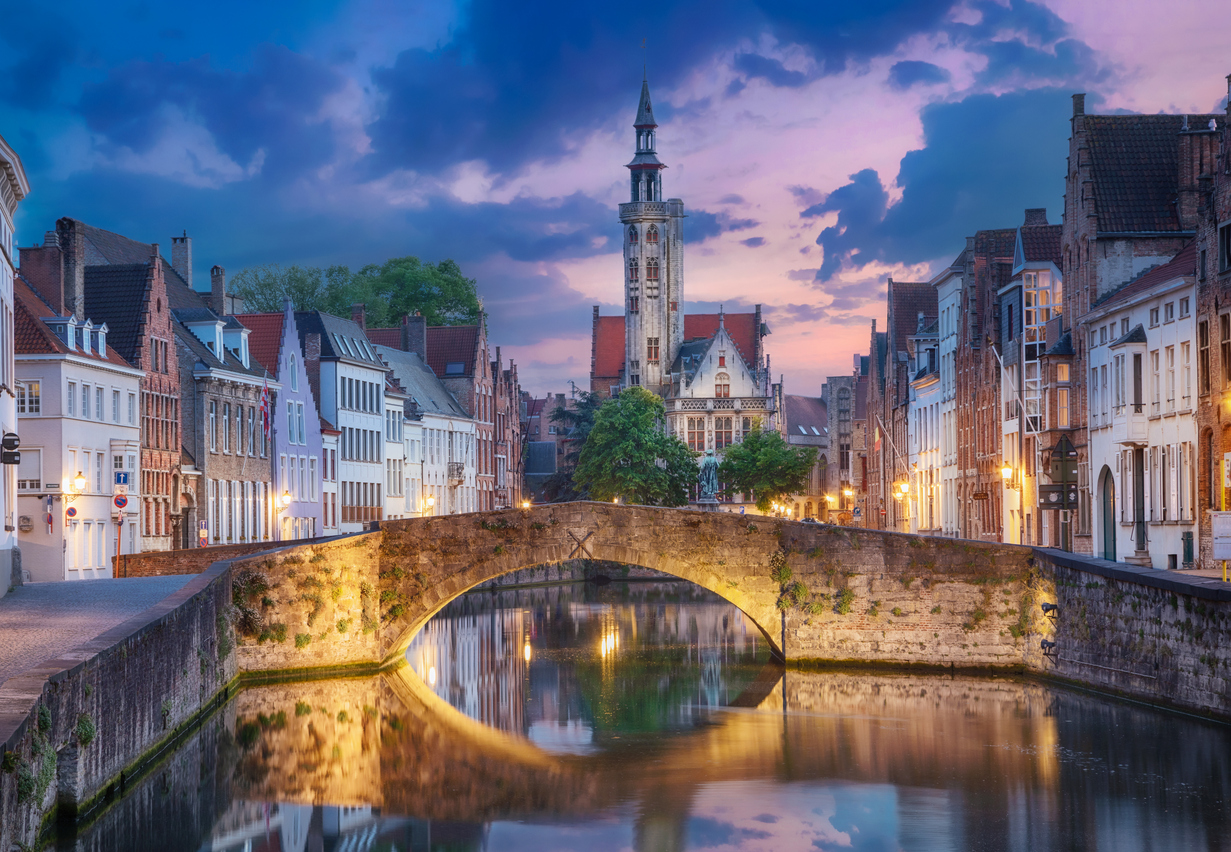 Beleuchtete Steinbrücke, historische Giebelhäuser und ein Turm spiegeln sich im ruhigen Kanal von Brügge zur Abenddämmerung.
