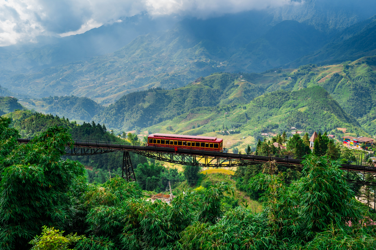 Roter Bergbahnwagen überquert einen schmalen Stahlviadukt vor grünen Bergen in Sapa, Vietnam.