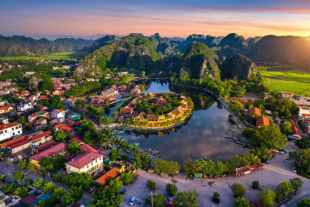 Luftaufnahme der Ninh-Binh-Karstlandschaft mit kreisförmiger Inselanlage in einem See, umgeben von grünen Bergen und Häusern.