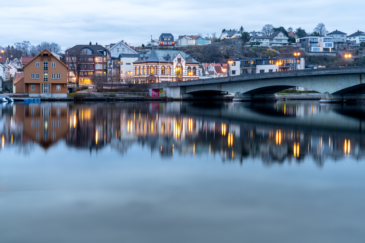 Blick über die Otra in Kristiansand (Sørlandet) mit beleuchteter Brücke, Uferhäusern und ruhigen Wasserspiegelungen in der blauen Stunde.